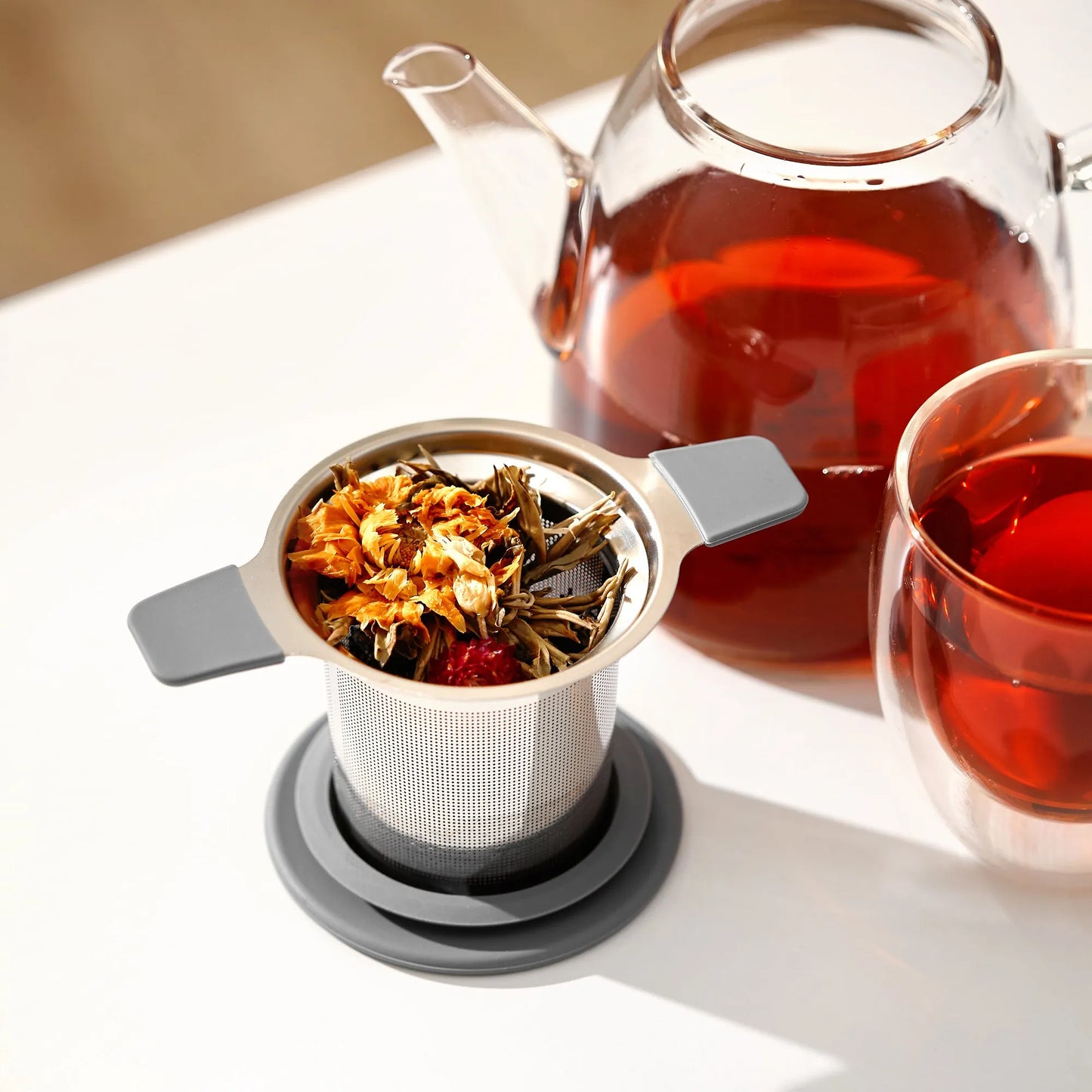 Tea infuser with loose tea leaves next to a glass teapot and cup filled with tea on a white surface.
