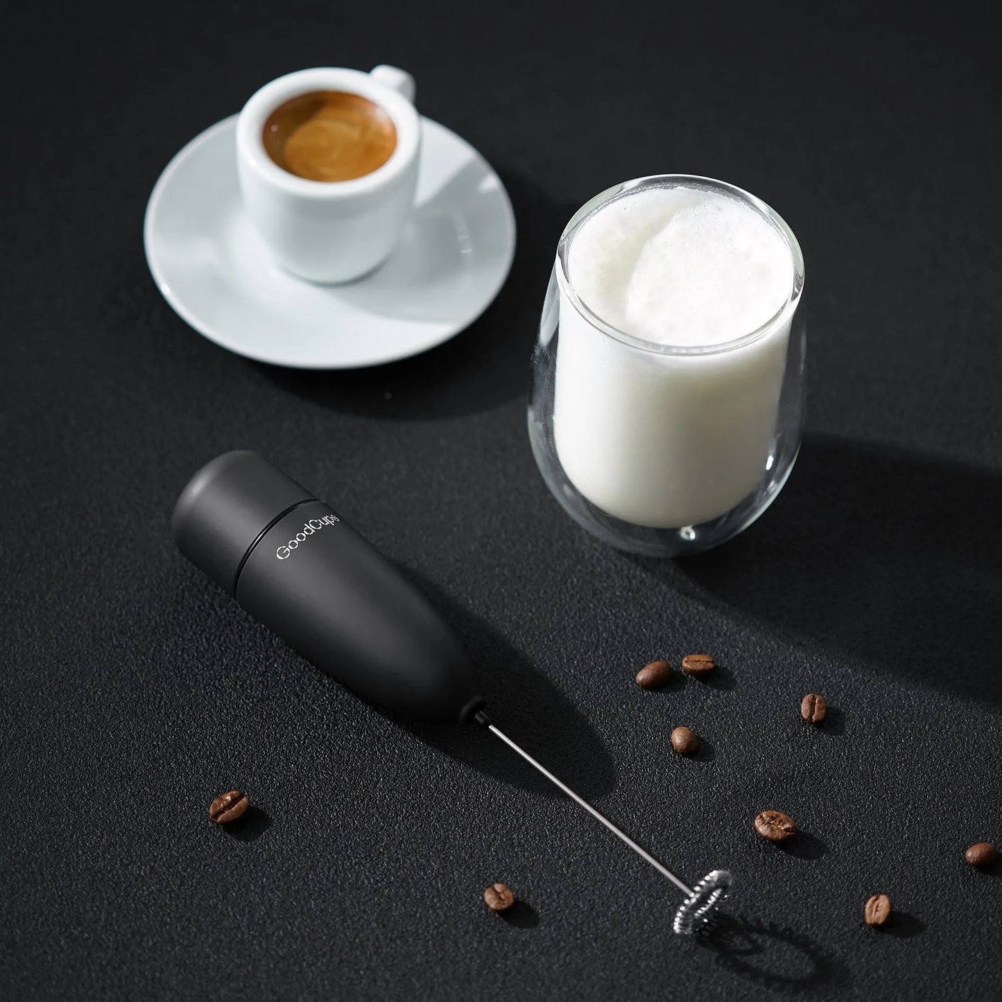 Black milk frother with a cup of coffee and glass of milk on a dark surface.