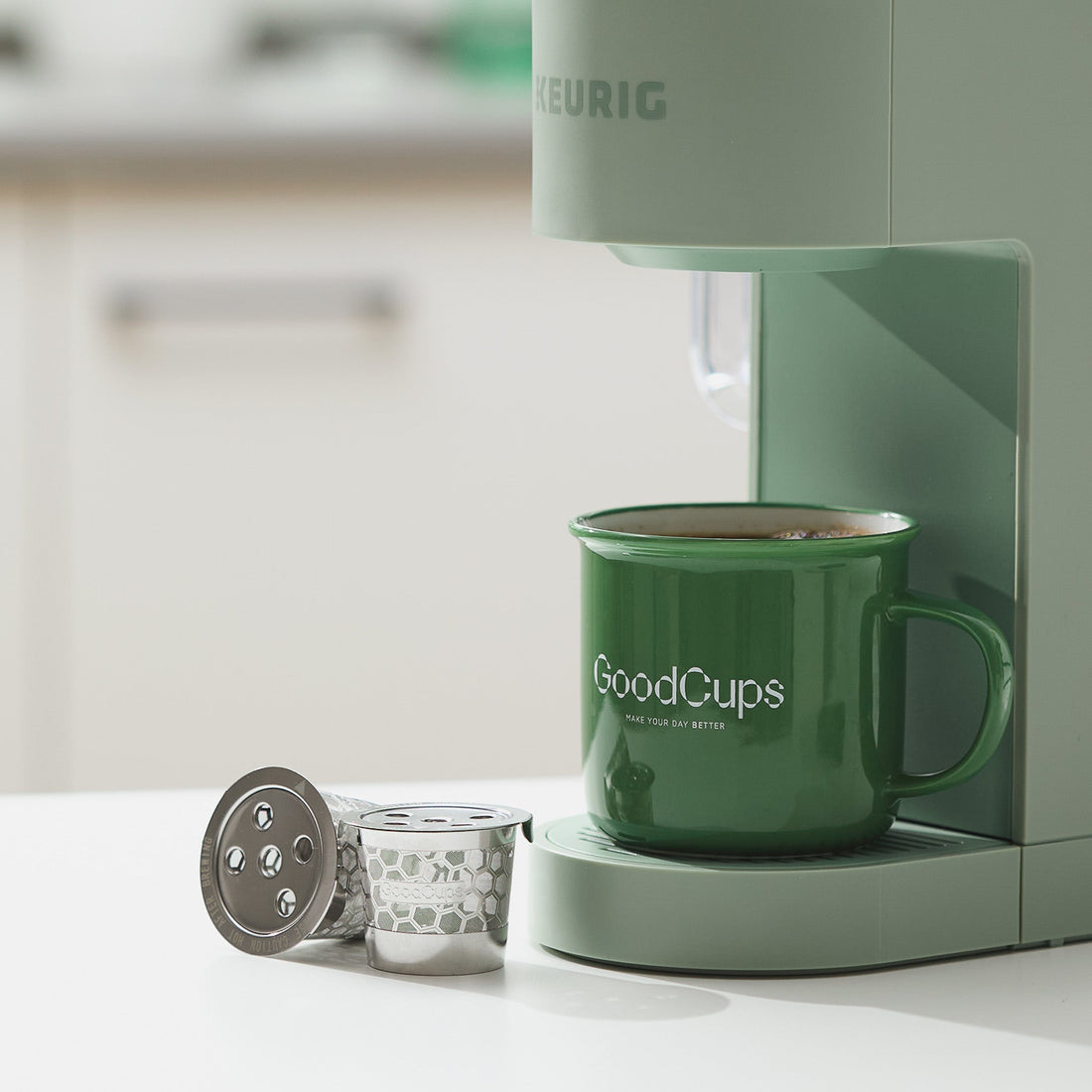 GoodCups stainless steel reusable K-Cups next to a Keurig coffee maker and GoodCups mug