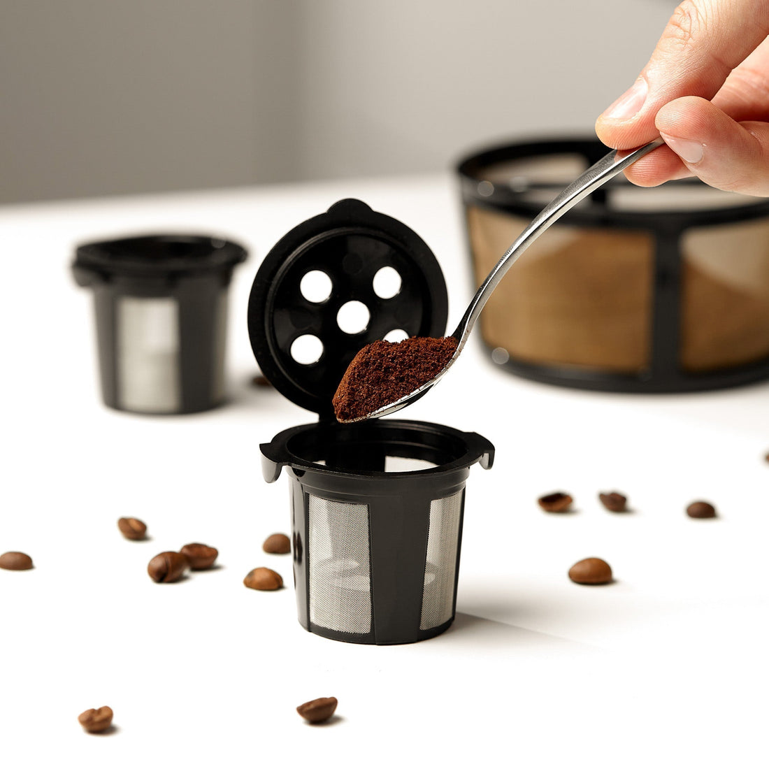 Spoon adding ground coffee into a GoodCups reusable K-Cup, with a MultiStream pod and coffee beans nearby
