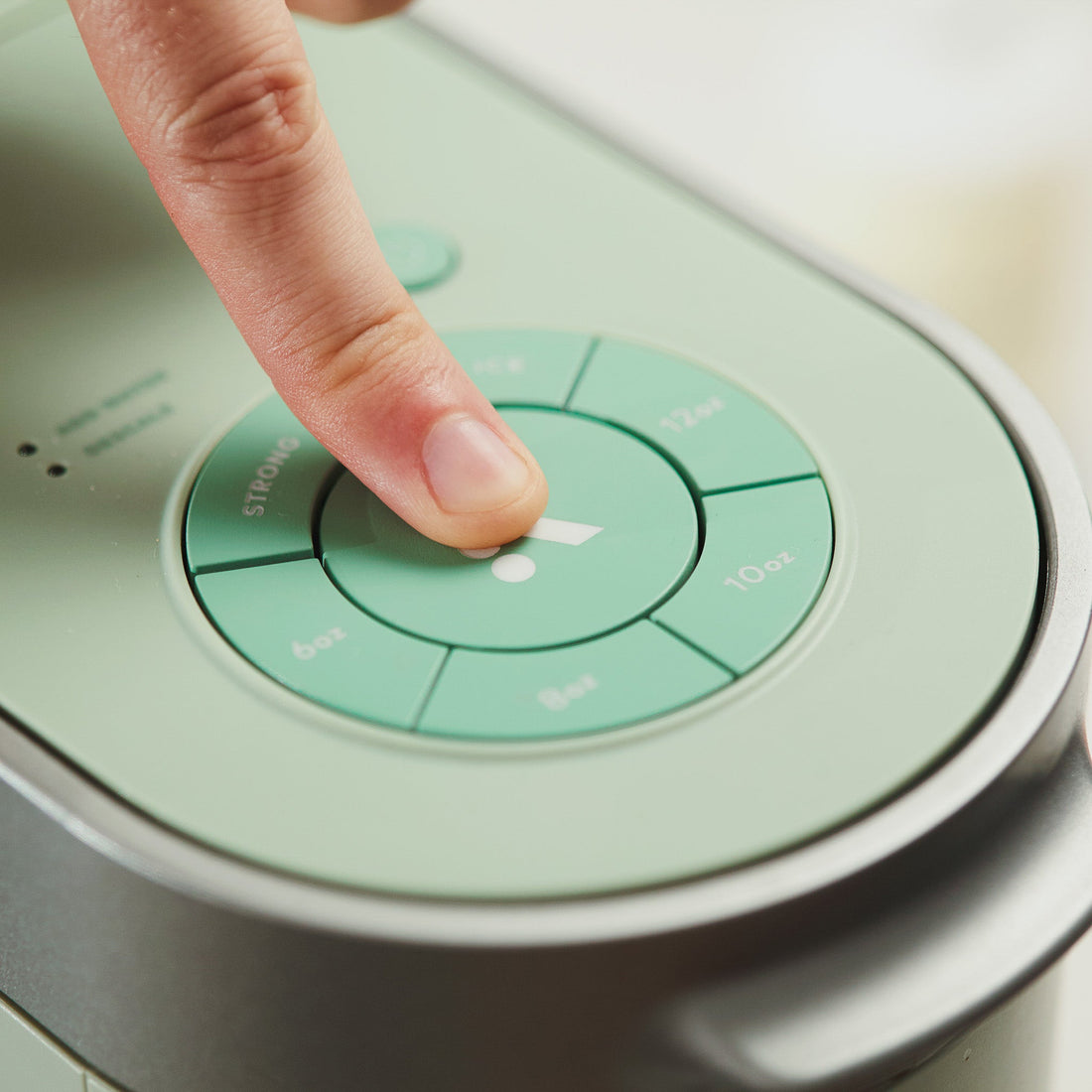 Finger pressing a brew-size button on the control panel of a mint green Keurig brewer, with STRONG, 6oz, 8oz, 10oz and 12oz options visible