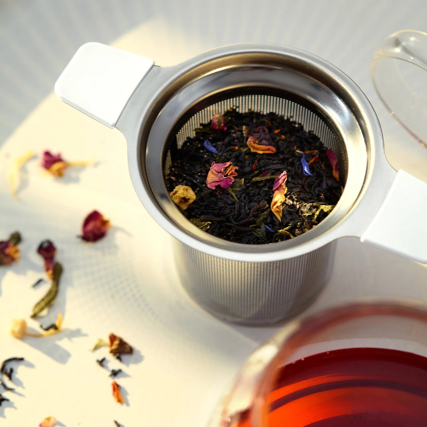 Tea strainer with dried tea leaves and flowers on a light surface, with a glass of tea in the background.