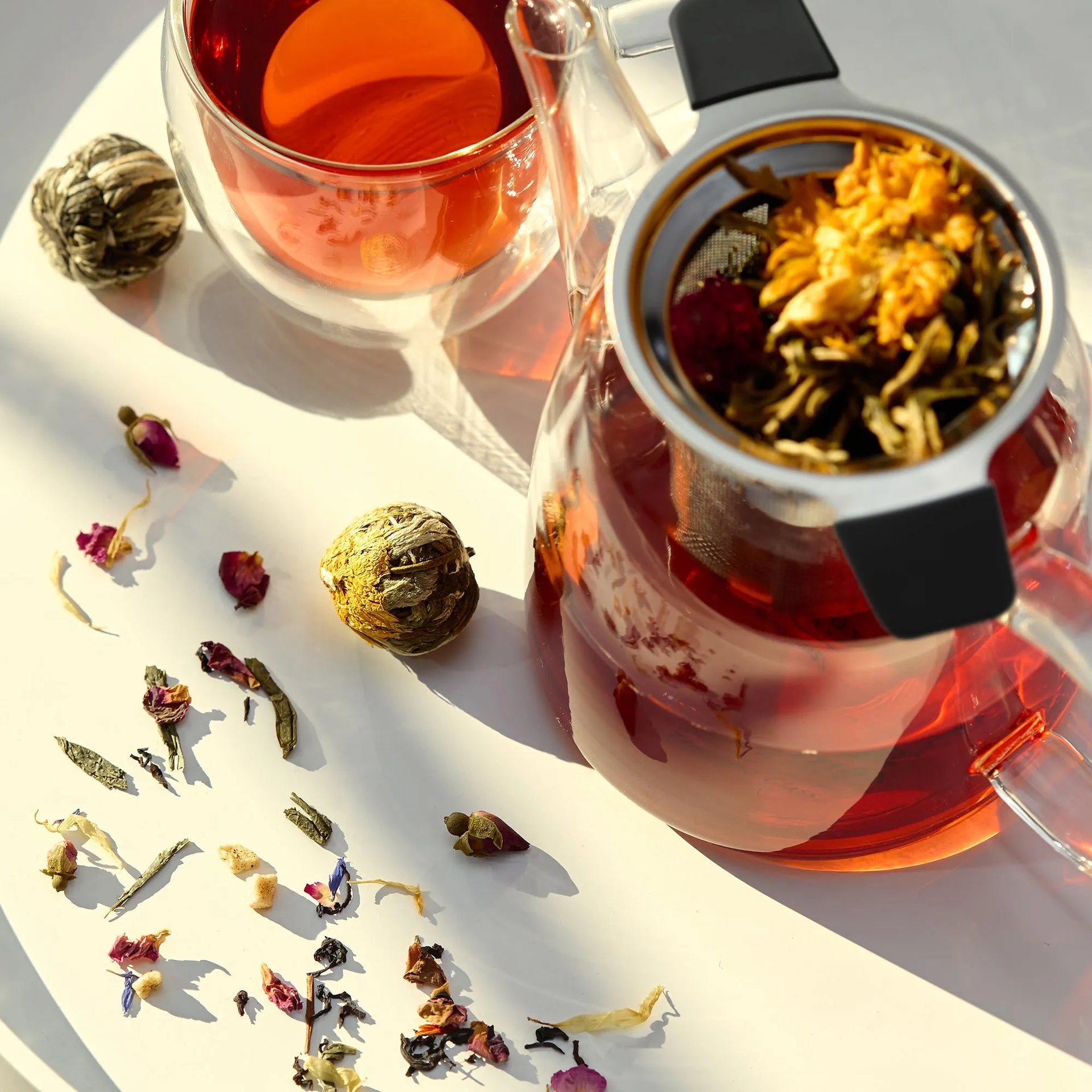 Tea-making setup with a glass teapot, cup, and various tea leaves on a white surface.