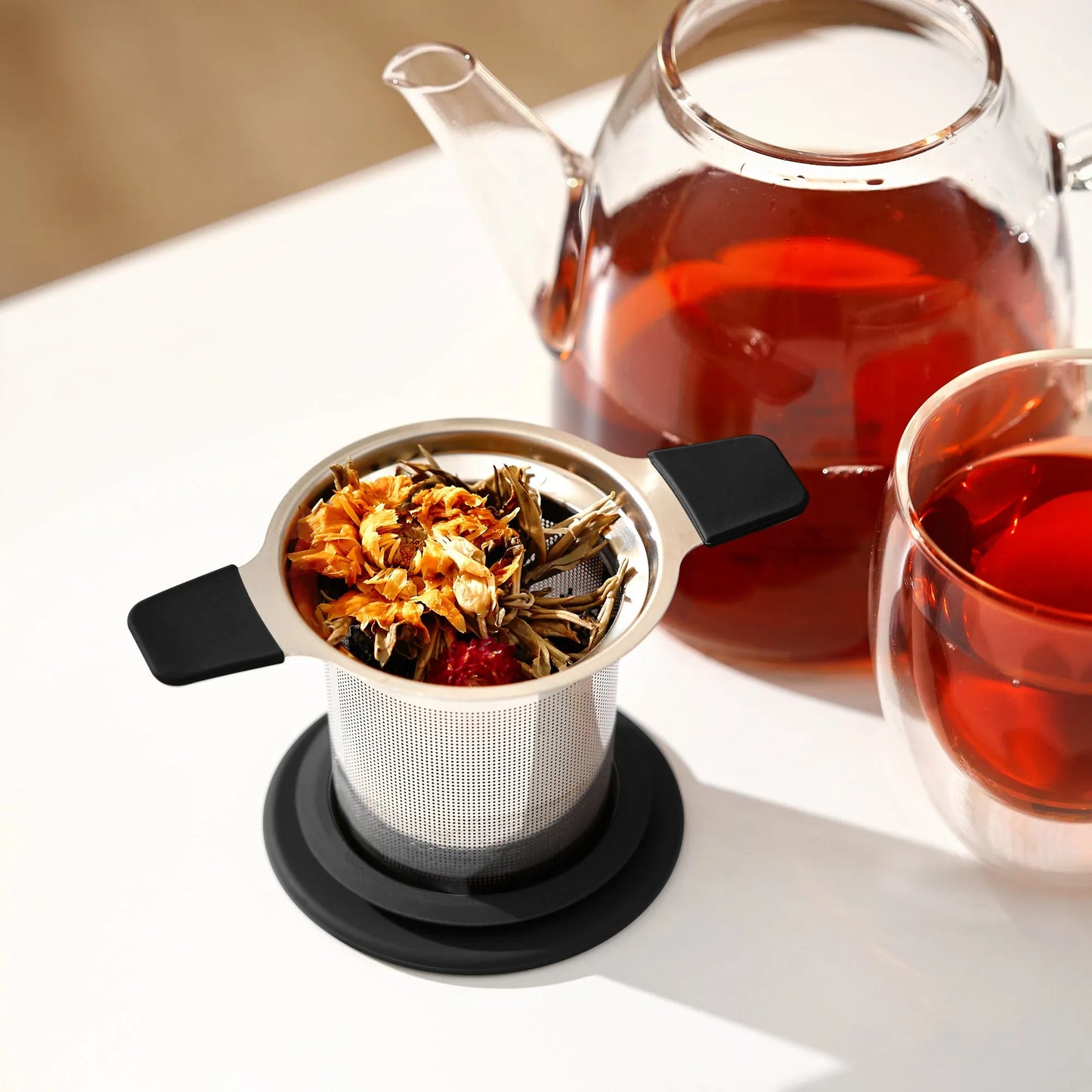 Tea infuser with loose tea leaves, glass teapot, and cup on a white surface