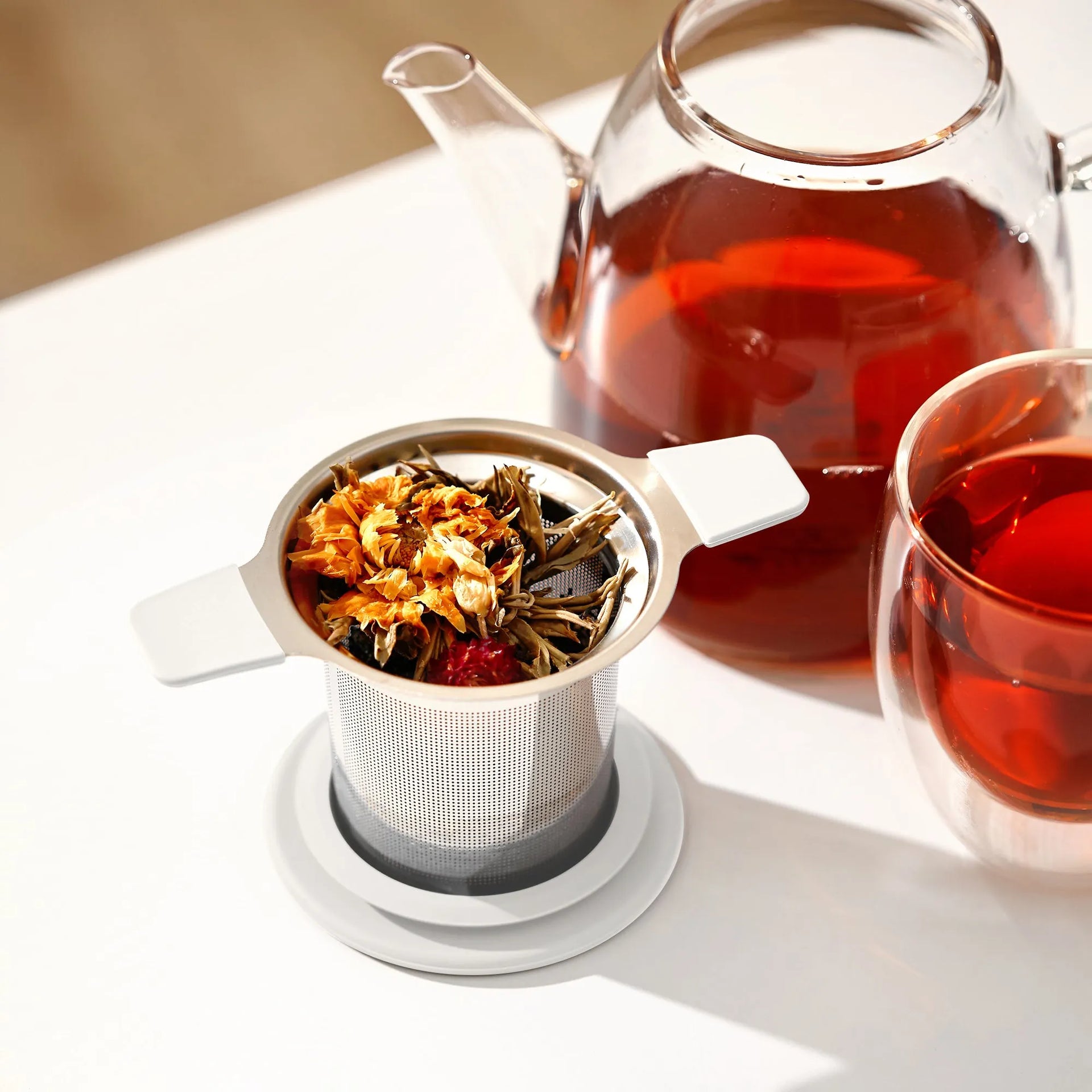 Tea infuser with loose tea leaves, glass teapot, and cup of tea on a white surface.