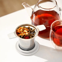Tea infuser with loose tea leaves, glass teapot, and cup of tea on a white surface.