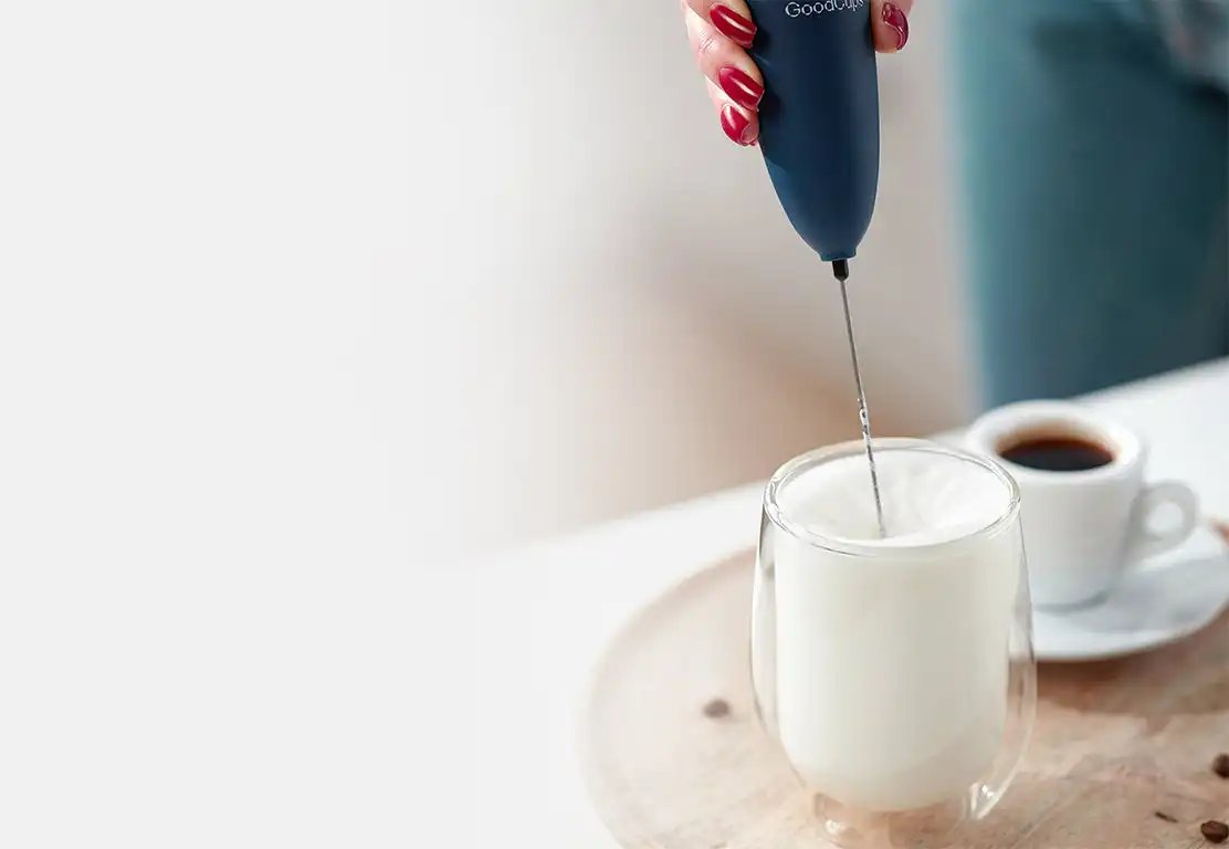 Hand using a milk frother to froth milk in a glass with a blurred background.