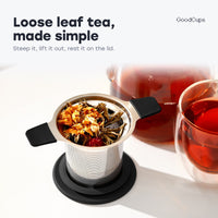 Tea infuser with black handles and loose leaf tea, resting on its lid by a glass teapot
