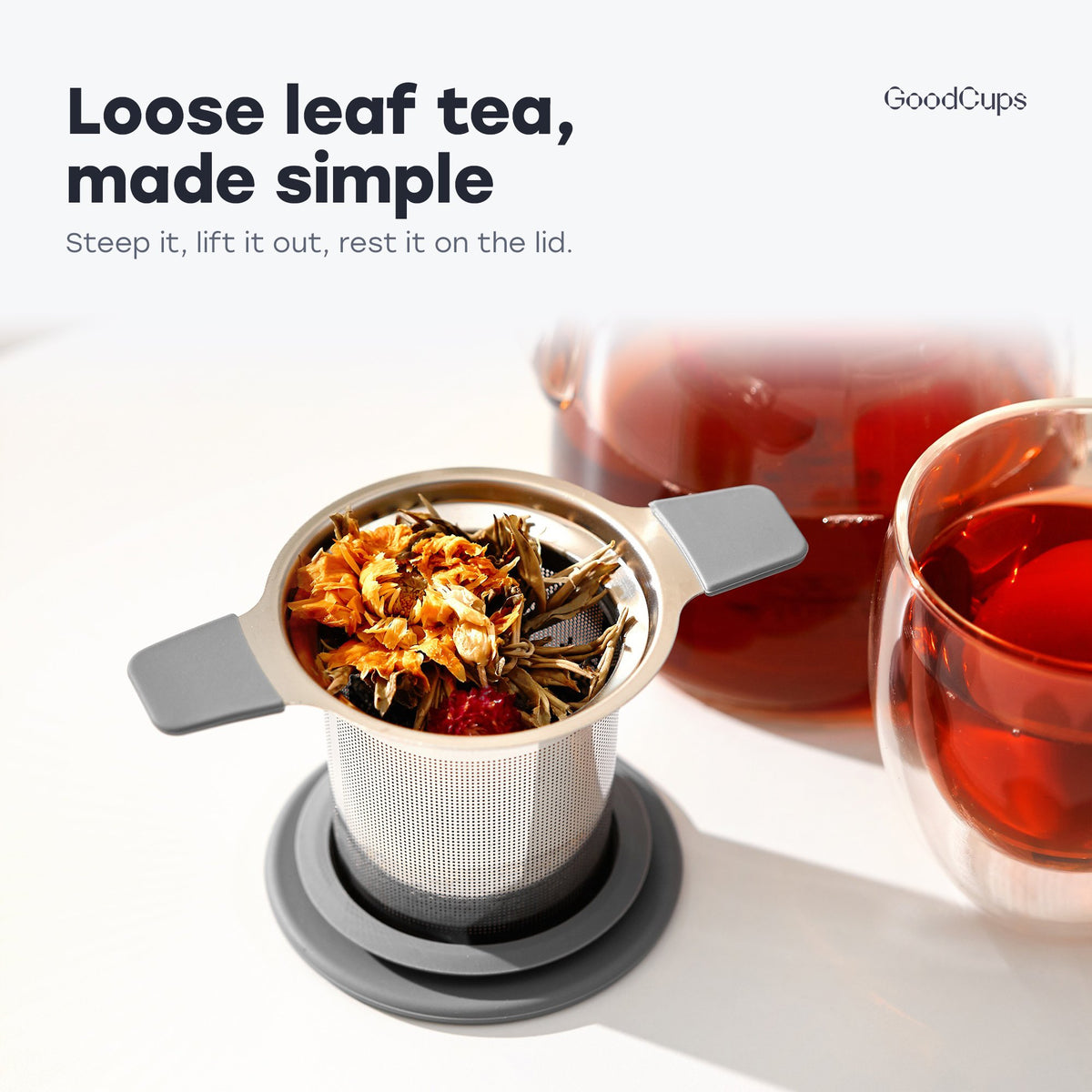 Tea infuser with gray handles and loose leaf tea, resting on its lid by a glass teapot