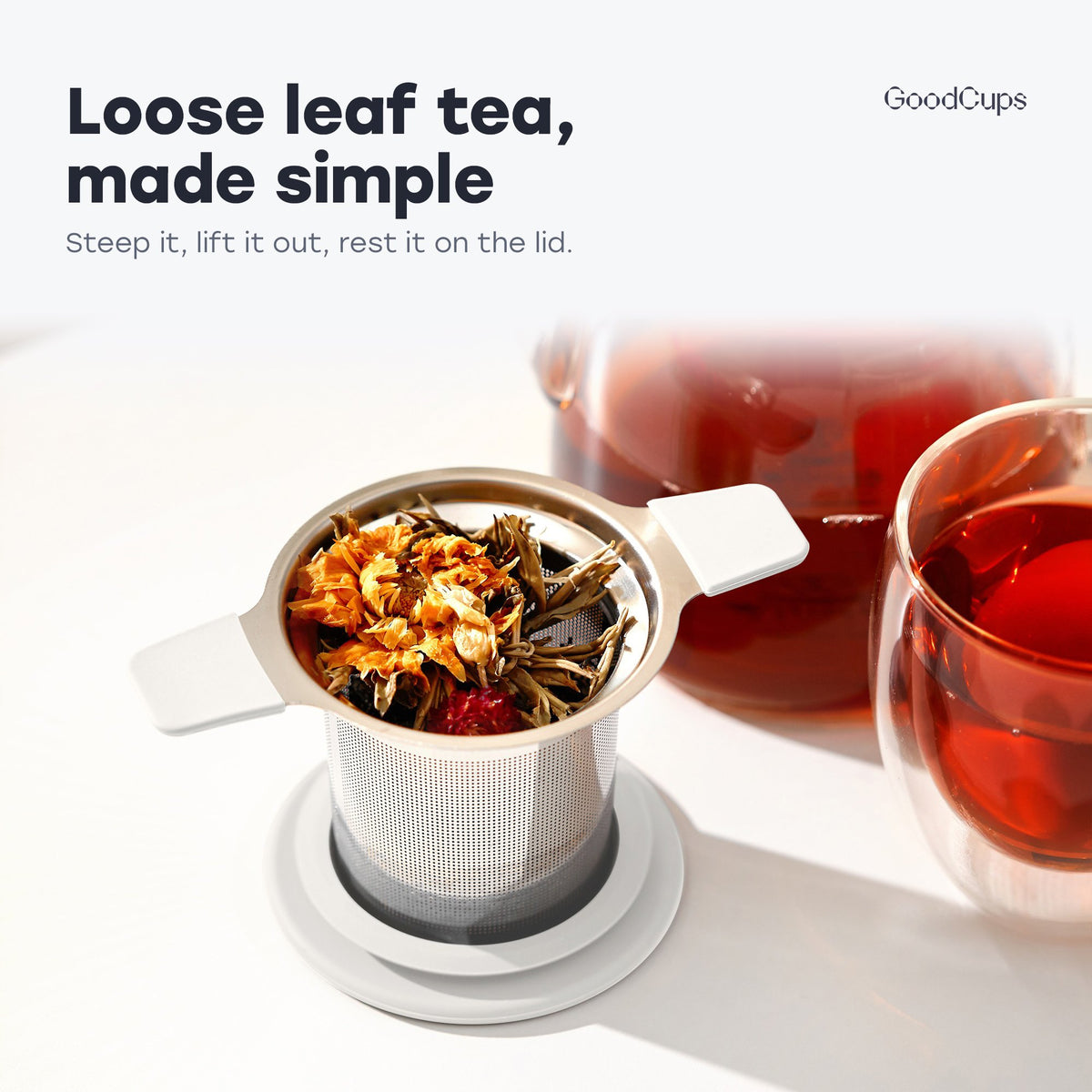 Tea infuser with white handles and loose leaf tea, resting on its lid by a glass teapot