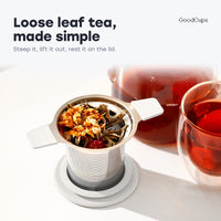 Tea infuser with white handles and loose leaf tea, resting on its lid by a glass teapot