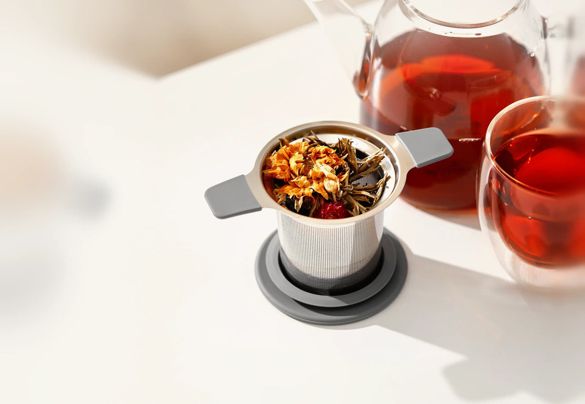 GoodCups stainless steel tea infuser with handles and loose leaf tea next to a glass teapot